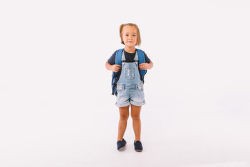 Little girl with blond hair dressed in a blue dungarees and t-shirt, with a backpack ready for back to school, on white background