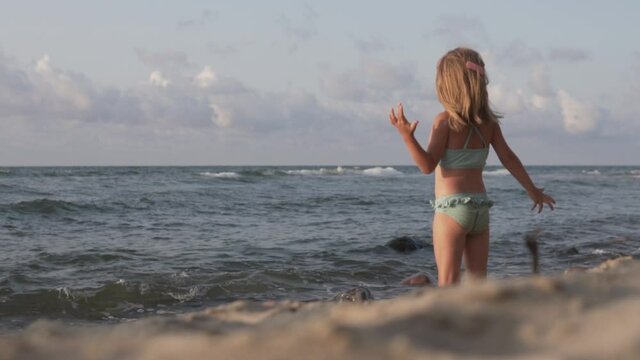 A beautiful little girl in a swimsuit stands on the beach by the sea, the sand in the foreground is blurred.