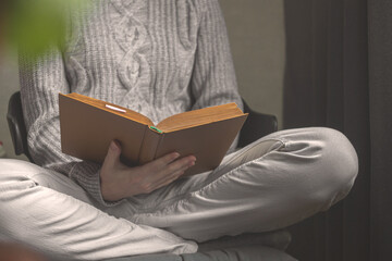 Elderly woman in woolen sweater reading old book sitting on chair at home. Cozy indoors holidays concept background photo