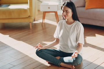 Asian woman doing yoga and zen like meditation in lotus pose in casual wear at indoor living room...