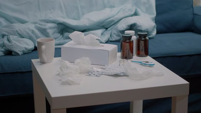 Close up of table with medicaments, thermometer and tissues in empty room. Nobody in space with medical treatment, tablets of capsules, bottles of pills and cup of tea for ill person