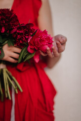 Lady in the red gorgeous dress gently touching pink bouquet of peony above white studio backdrop. Female came after the date in a romantic mood with her favourite flowers which smell so magnificent 