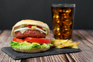 Hamburguesa vegana vegetal con queso sobre un plato negro de pizarra, con patatas fritas y un refresco de fondo, sobre una mesa de madera y un fondo negro
