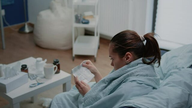 Sick adult reading medicament information on paper to heal virus infection. Woman feeling cold and holding package leaflet while sitting in blanket for seasonal flu treatment.
