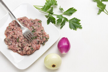Minced meat and fork on plate. Peeled onions and sprig of parsley on table