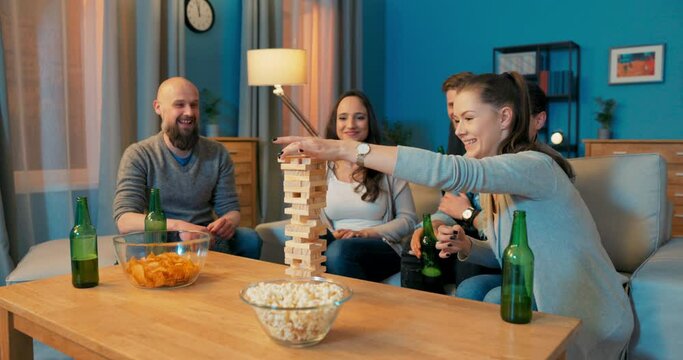 People playing game in evening, sharing chips, drinking beer, taking turns pulling out pieces of tower woman grabs block quickly pulls it out causing collapse of structure, grabs head covers legs
