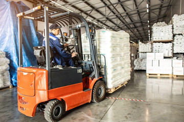 A man on a forklift works in a large warehouse, unloads bags of raw materials - obrazy, fototapety, plakaty