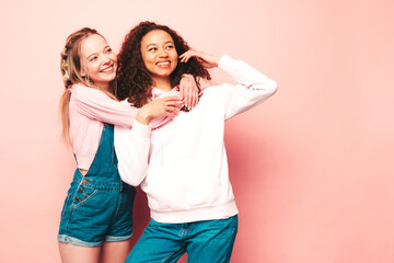 Two young beautiful smiling international hipster female in trendy summer clothes. Sexy carefree women posing near pink wall in studio. Positive models having fun. Concept of friendship