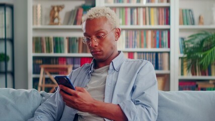 Confident and serious dark-skinned man resting sitting on sofa and typing on smartphone. Portrait of stylish young man in casual clothes checking social networks sitting at home in living room.