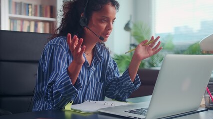 Young female operator in headphones talking on phone sitting at her workplace in front of a laptop. Indian young woman looking at laptop screen having a pleasant conversation with clients or partners.