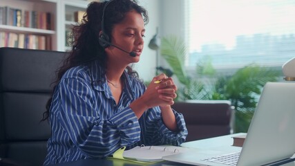 Young Indian female student in headset discusses test result with tutor at online conference. Focused woman in headphones sitting in front of laptop and learning foreign language on distant course.