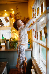 Toddler child hangs holiday flags on the window with garlands and lights, festive mood. Birthday and Christmas concept, holiday preparation