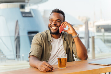 Portrait of a young black man talking to his mobile phone while sitting with a beer and a laptop in a bar.