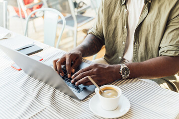 Close up view of the hands of a young african man typing on a laptop in a coffee shop