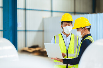 Team engineers and foreman wear a mask, hard hat, and vest. Standing consult discuss industrial production management. Explaining job details through laptops and tablets in factories or warehouses.