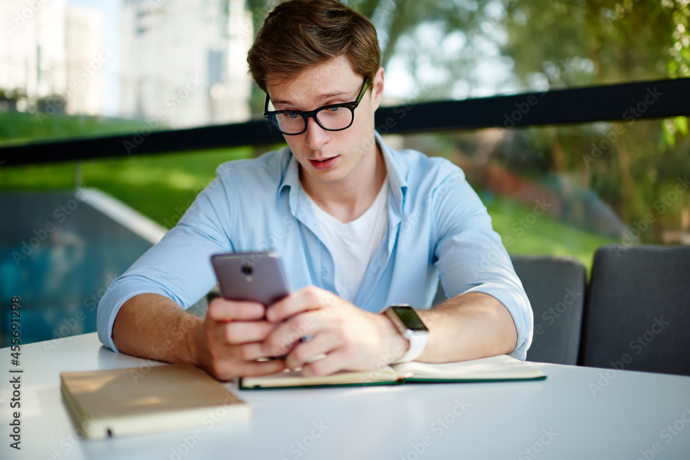Poster caucasian male student in optical eyeglasses reading shocked news while networking social media via 