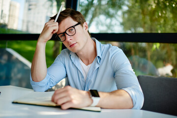 Portrait of male student in optical spectacles for provide eyes correction looking at camera while thinking about education learning in cafe interior, pensive man in glasses studying indoors