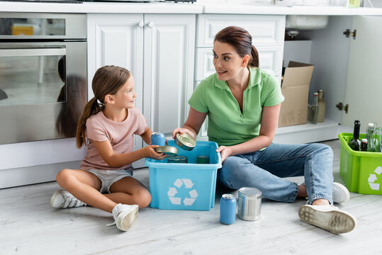 Positive girl putting tin cans in box with recycle sign near mother in kitchen
