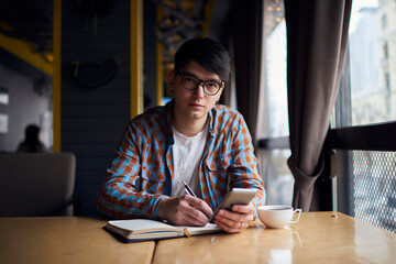 Millennial mobile phone user sitting at the table with notebook while preparing for exams. Smartphone application, modern technology for learning