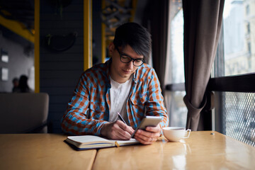 Millennial mobile phone user sitting at the table with notebook while preparing for exams. Smartphone application, modern technology for learning