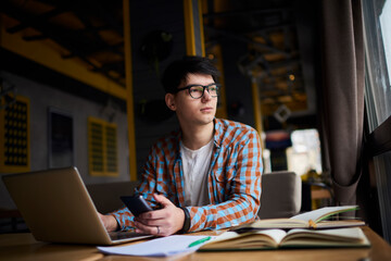 Fototapeta premium Millennial male using mobile phone while sitting at the table with books while front open laptop computer. Smartphone application. Working on freelance, using technology