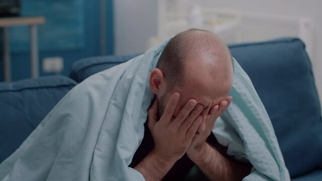 Close up of sick person rubbing temples against headache feeling miserable because of disease. Unwell man with migraine and virus symptoms while holding hands on head in blanket.
