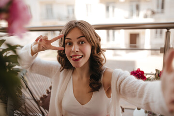 Close up shot of young latvian lady showing peace sign and smiling widely. Sitting on the veranda of provance house in milky singlet and vintage snowy cardigan with pink lipstick and beautiful light 