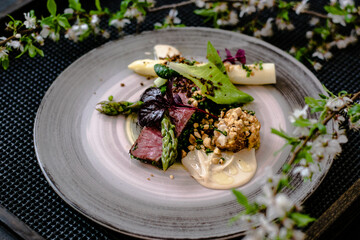Dish of ostrich steak with white and green asparagus, garden greens and sauce on a plate on wooden table in a restaurant