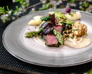Foto op Plexiglas Dish of ostrich steak with white and green asparagus, garden greens and sauce on a plate on wooden table in a restaurant © Mzals
