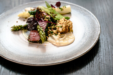 Dish of ostrich steak with white and green asparagus, garden greens and sauce on a plate on wooden table in a restaurant
