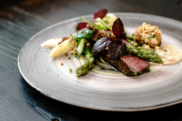 Dish of ostrich steak with white and green asparagus, garden greens and sauce on a plate on wooden...