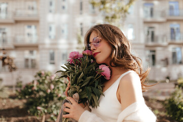 Inspired french madame tilted her head and touches softly bouquet of flowers. Dark haired girl with nude make up, red lipstick and snowy cardigan posing outdoors in the golden hour