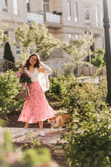 Obraz na płótnie Canvas Lady posing full size in the beautiful garden during sunny spring day. Female in flying soft raspberry dress and short brown hair holds bouquet of peonies and walking with dog