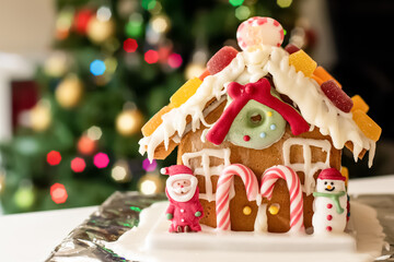 Homemade gingerbread house decorated with icing, sweets , figurines, lollies and candy canes with blurred Christmas tree on background