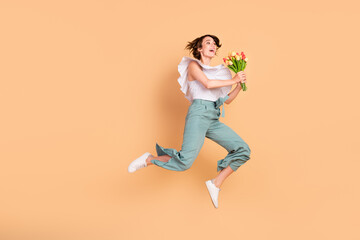 Fototapeta na wymiar Photo of funny impressed young woman dressed striped top jumping high holding flower blossom isolated beige color background