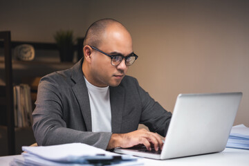 Young asian businessman working with laptop computer in the dark office at night. Attractive Indian man work hard overtime serious thinking in home with floor lamp ambient warm light late at night