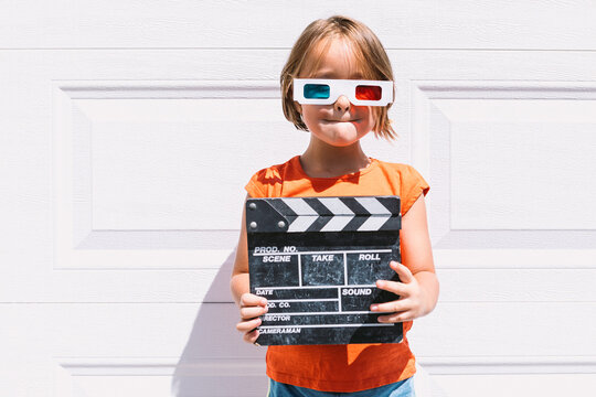 Trendy kid in 3D glasses holding clapperboard standing on wall