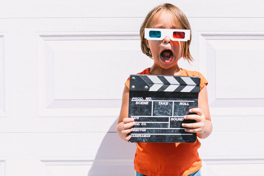 Trendy kid in 3D glasses holding clapperboard standing on wall