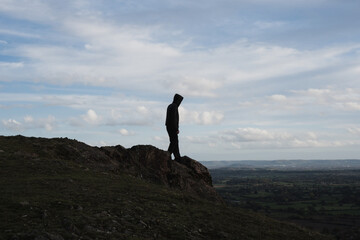 A hooded figure walking towards the top of a rocky outcrop. Looking down across the landscape