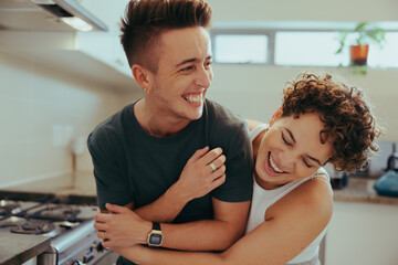 Young queer couple laughing cheerfully in their kitchen - obrazy, fototapety, plakaty