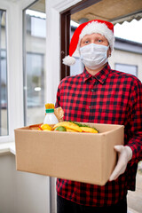 Volunteer man in Santa hat and protective mask and gloves delivery donation box at home in Christmas