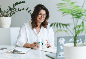 Young smiling brunette woman nutritionist plus size in white shirt working at laptop on table with...