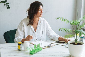 Young smiling brunette woman nutritionist plus size in white shirt working at laptop on table with house plant in bright modern office