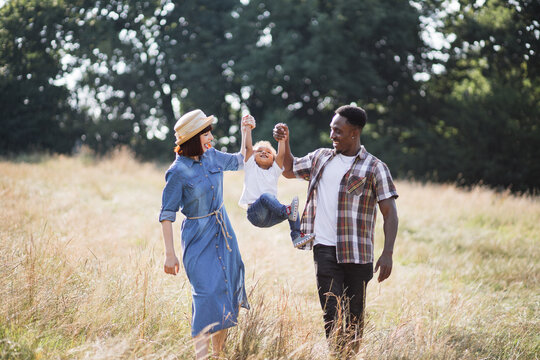Happy multi ethnic parents playing with son while raising him by arms outdoors. Caucasian woman and african man enjoying time with little child on fresh air.
