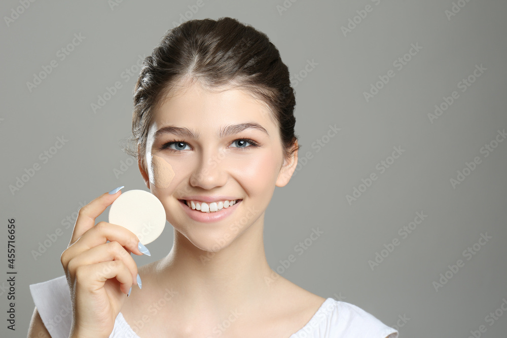 Wall mural Beautiful girl with foundation smear on her face holding sponge against grey background