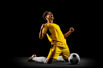 Full-length horizontal portrait of young man, male soccer football player training isolated on black background.