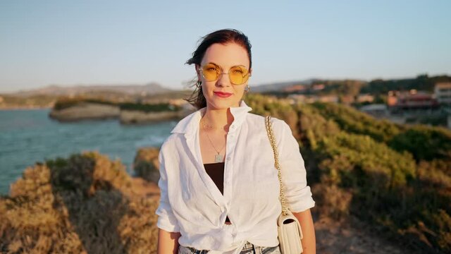 Portrait of woman in sunglasses smiling, she standing on cliff beach. Blue sea background. Lady in white linen shirt. Travel destination, vacation in Greece.