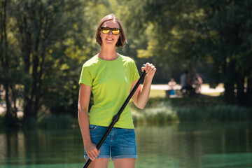 Smiling young woman on SUP with sunglasses holding a paddle, in the background visible lake water and green trees
