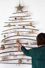 Back view of woman decorating handmade craft Christmas tree made from sticks and natural materials hanging on wall.