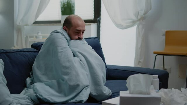 Person with flu feeling cold and shivering wrapped in blanket. Sick man having chills and fever symptoms while trying to get warm with cover, to cure illness with medicaments on table.